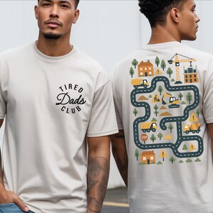May include: Beige t-shirt with the text "Tired Dads Club" on the front. The back of the shirt features a colorful construction-themed graphic with trucks, a crane, houses, and roads.