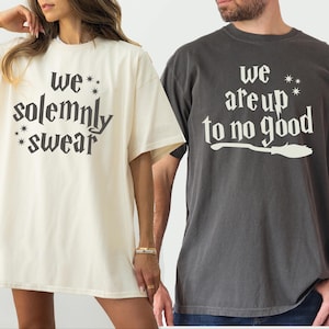 May include: Two oversized t-shirts, one cream and one charcoal gray. The cream shirt reads "we solemnly swear" in a stylized font. The gray shirt says "we are up to no good" with a broom graphic. Both shirts are short-sleeved.