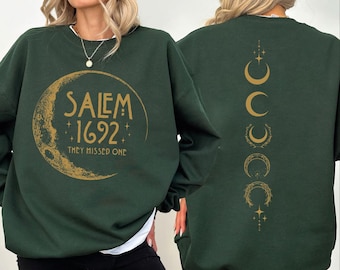 May include: Dark green sweatshirt with gold text and graphics. The front features a crescent moon with the words "Salem 1692" and "They Missed One." The back has a vertical design of crescent moons. The sweatshirt has a crew neck and long sleeves.