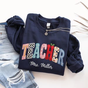 May include: A navy blue crewneck sweatshirt featuring the word TEACHER in large, multi-patterned embroidered letters, with the name Mrs. Miller embroidered in white script below. The sweatshirt is paired with distressed light blue denim jeans.
