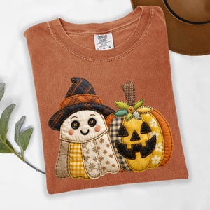May include: A rust-colored t-shirt featuring a graphic of a ghost wearing a plaid witch hat and a jack-o'-lantern pumpkin. The ghost has button eyes and a stitched smile. The pumpkin has a checkered pattern and a small green fabric leaf accent.