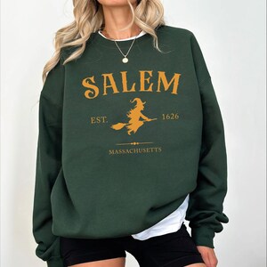 A woman wearing a green sweatshirt with the text "SALEM EST. 1626 MASSACHUSETTS" printed on it.