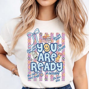 May include: Cream-colored t-shirt with a colorful graphic. The design features the words "YOU ARE READY" in blue, a smiley face, and supporting text like "Show what you know" and "Do your best". The background has a floral pattern.