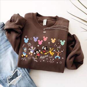 A brown sweatshirt with a Disney character design, including Mickey Mouse, Minnie Mouse, Donald Duck, Goofy, and Pluto, along with various Disney-themed elements such as stars, hearts, and balloons.