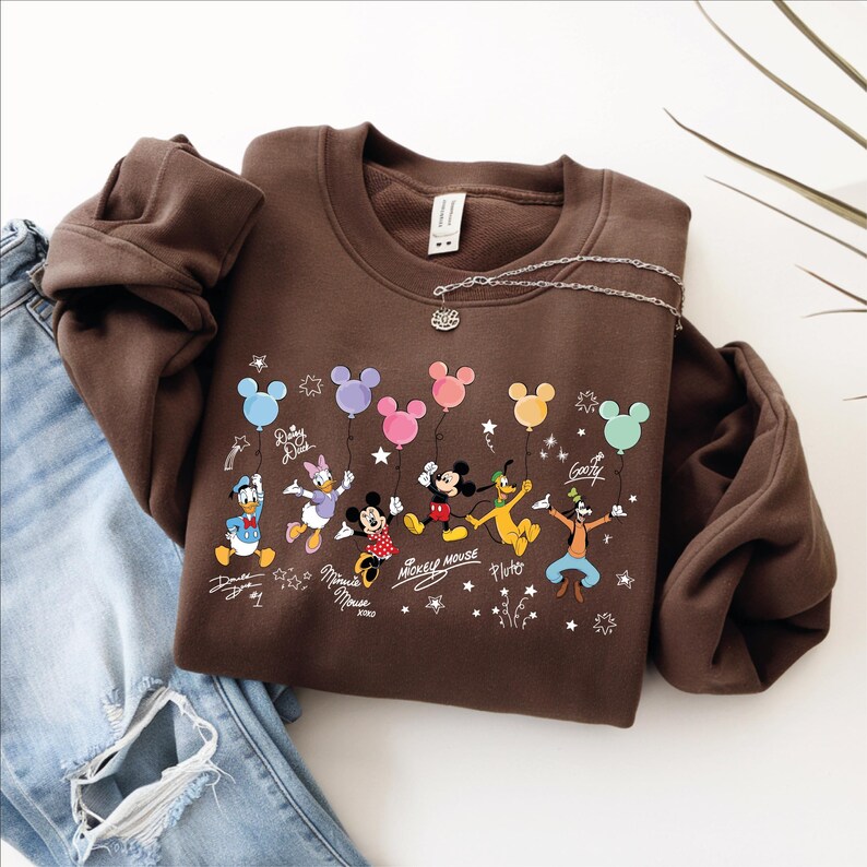 A brown sweatshirt with a Disney character design, including Mickey Mouse, Minnie Mouse, Donald Duck, Goofy, and Pluto, along with various Disney-themed elements such as stars, hearts, and balloons.