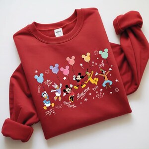 A red sweater with a Disney character design, including Mickey Mouse, Minnie Mouse, Donald Duck, Goofy, and Pluto, along with various Disney-themed elements such as balloons and stars.