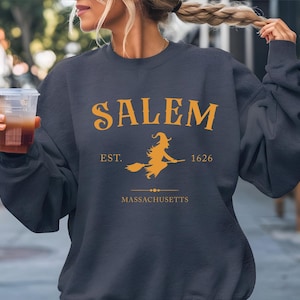 A woman wearing a navy blue sweatshirt with the text "SALEM EST. 1626 MASSACHUSETTS" printed on it, holding a drink in her hand.