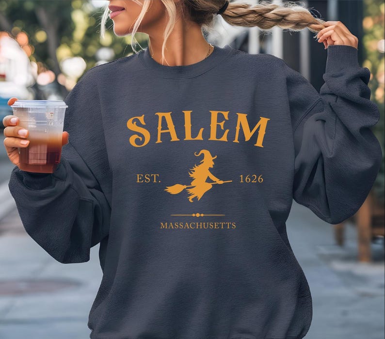 A woman wearing a navy blue sweatshirt with the text "SALEM EST. 1626 MASSACHUSETTS" printed on it, holding a drink in her hand.