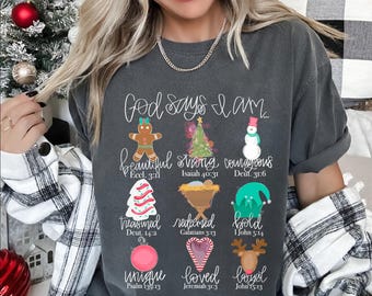 Puede incluir: Camiseta gris oscuro con el texto "Dios dice que soy" e ilustraciones navideñas. La camiseta presenta un hombre de jengibre, un árbol de Navidad, un muñeco de nieve y otros diseños festivos. También están impresas las palabras "hermoso, fuerte, valiente, atesorado, redimido, audaz, único, amado, leal".