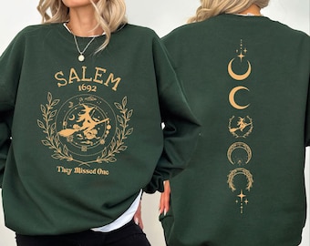 May include: Dark green sweatshirt with gold-colored Salem 1692 graphic on the front, featuring a witch on a broom. The back displays a moon phase design with a witch silhouette. The text "They Missed One" is below the front graphic.