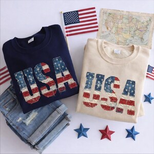 May include: Two crewneck sweatshirts, one navy blue and one beige, each with "USA" in a distressed American flag design. A pair of rolled-up denim shorts, miniature American flags, a vintage map, and star-shaped decorations complete the patriotic theme.