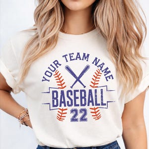 May include: Cream-colored baseball t-shirt with a blue and red graphic. The graphic includes crossed baseball bats, the word "BASEBALL", the number "22", and space for a team name.