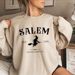 A woman wearing a beige sweatshirt with the text "SALEM EST. 1626 MASSACHUSETTS" printed on it.