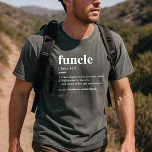 May include: Dark green t-shirt with the word "funcle" in white, along with a definition. The definition includes the pronunciation, and describes a funcle as a cooler uncle, well-trained in arts and amusement. The shirt is worn by a person with a backpack.