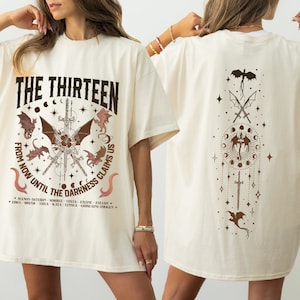 May include: A white oversized t-shirt with a graphic print of a circle with the text "THE THIRTEEN" and a design of swords and dragons. The text below the circle reads "FROM NOW UNTIL THE DARKNESS CLAIMS US".