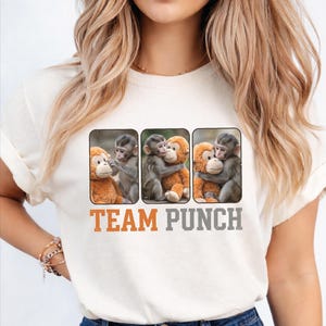 May include: Cream-colored t-shirt featuring three rectangular images of monkeys hugging stuffed animals. The text "TEAM PUNCH" is printed in orange and gray below the images. The shirt is made of a soft material.