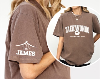 May include: A brown t-shirt with the words "TAEKWONDO mom" in white lettering. The shirt also features a white graphic on the left sleeve with the name "JAMES" below it. The shirt is a comfortable, casual style.