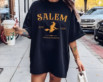 May include: Black t-shirt with the word "SALEM" in gold lettering, along with a witch on a broom and the text "EST. 1626 MASSACHUSETTS". The shirt is oversized and has short sleeves. The person is holding a white bag and a coffee cup.