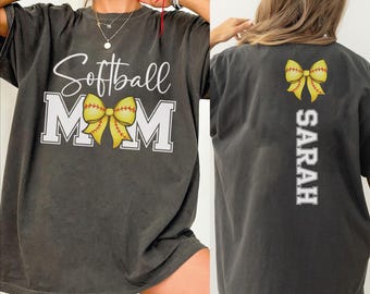 May include: Dark gray t-shirt with "Softball Mom" in white script and block letters, featuring a yellow softball bow. The back of the shirt has a matching bow and the name "Sarah" in white.