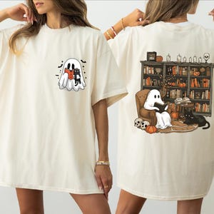 two women wearing white t-shirts with Halloween-themed designs. The t-shirt on the left features a ghost and a witch, while the t-shirt on the right depicts a ghost reading a book and a cat.