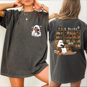 The image is a collage of two photos. The left photo shows a person wearing a gray t-shirt with a graphic of a ghost and a skeleton. The right photo shows the back of the same t-shirt, featuring a graphic of a ghost sitting in a chair surrounded by books and a skeleton.