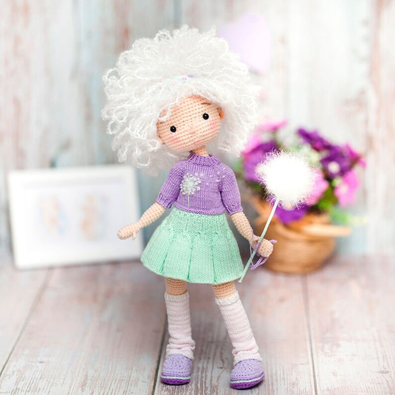 May include: A crocheted doll with white curly hair, a purple sweater with a flower design, a green skirt, and white socks. The doll is holding a white fluffy dandelion.