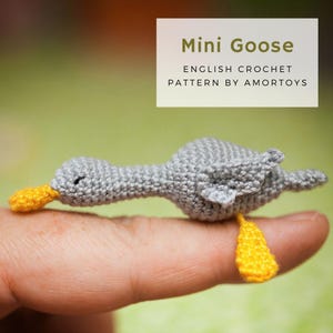May include: A small gray crocheted goose with yellow beak and feet. The goose is sitting on a finger.