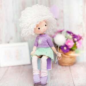 May include: A crocheted doll with white curly hair, wearing a purple sweater and a green skirt, holding a white dandelion. The doll is sitting on a stack of colorful boxes.