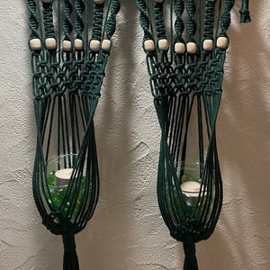 May include: A dark green macrame wall hanging with two hanging planters. The planters are made of rope and have wooden beads. The wall hanging is attached to a wooden branch.