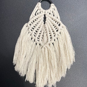 Può includere: Applique da parete in macramè color crema con un anello nero nella parte superiore. Il design presenta un motivo intrecciato con aperture a forma di diamante e lunghe frange nella parte inferiore. Questo pezzo fatto a mano aggiunge un tocco bohémien a qualsiasi spazio.