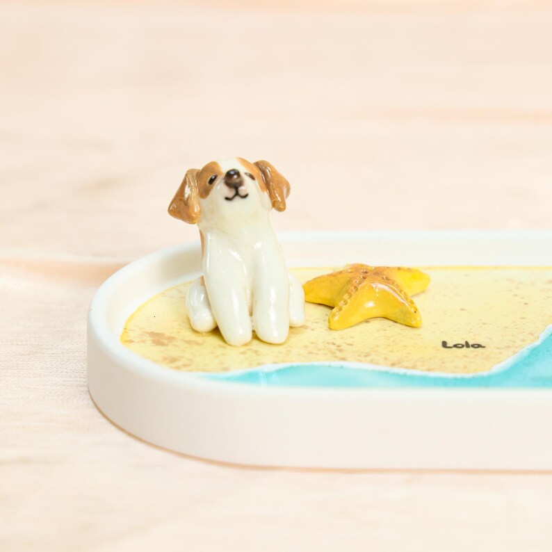 May include: A ceramic dog figurine with brown and white markings sits on a decorative tray. A yellow starfish and a beach scene are also depicted. The tray has a white rim and the word "Lola" is written on the tray.