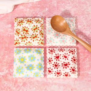 May include: Four square ceramic dishes with scalloped edges, each featuring a unique hand-painted pattern of red bows, blue flowers, or red hearts, accompanied by a single wooden spoon resting on the top right dish.