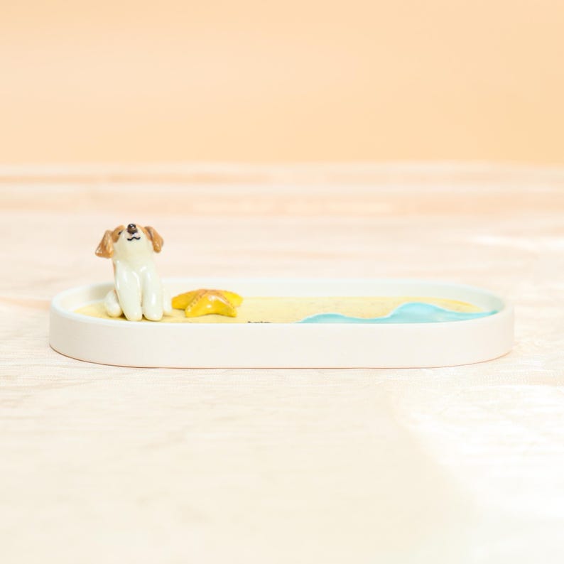 May include: A white ceramic tray featuring a beach scene. A small dog figurine in tan and white sits on the sand next to a yellow starfish. The tray has a light blue wave design, ideal for holding small items.