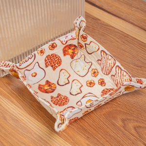 May include: A small, square, beige fabric tray with a white and orange pattern of various baked goods. The tray has four corners with gold metal snaps.