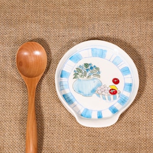May include: A wooden spoon lies next to a white ceramic spoon rest. The spoon rest has a blue and white striped border and a painted design of a blue vase with flowers and a bowl of fruit. The background is a textured tan fabric.