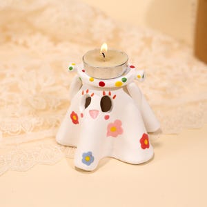 May include: A white ceramic candle holder shaped like a ghost, with a lit tea light candle on top. The ghost has a polka-dotted collar and is decorated with colorful flower accents. The candle holder is designed for decorative use.