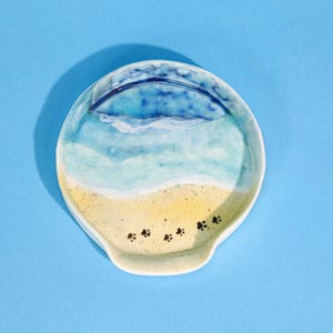 Puede incluir: Un reposacucharas de cerámica pintado a mano con una escena de playa. El diseño presenta un océano azul, olas blancas y una playa arenosa con huellas de patas negras. El reposacucharas es redondo con un pequeño mango.
