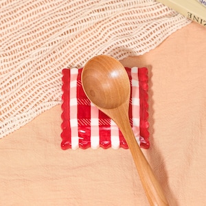 May include: A wooden spoon rests on a red and white checkered spoon rest. The spoon rest has a scalloped edge. The spoon and spoon rest are on a peach-colored surface with a white mesh cloth in the background.