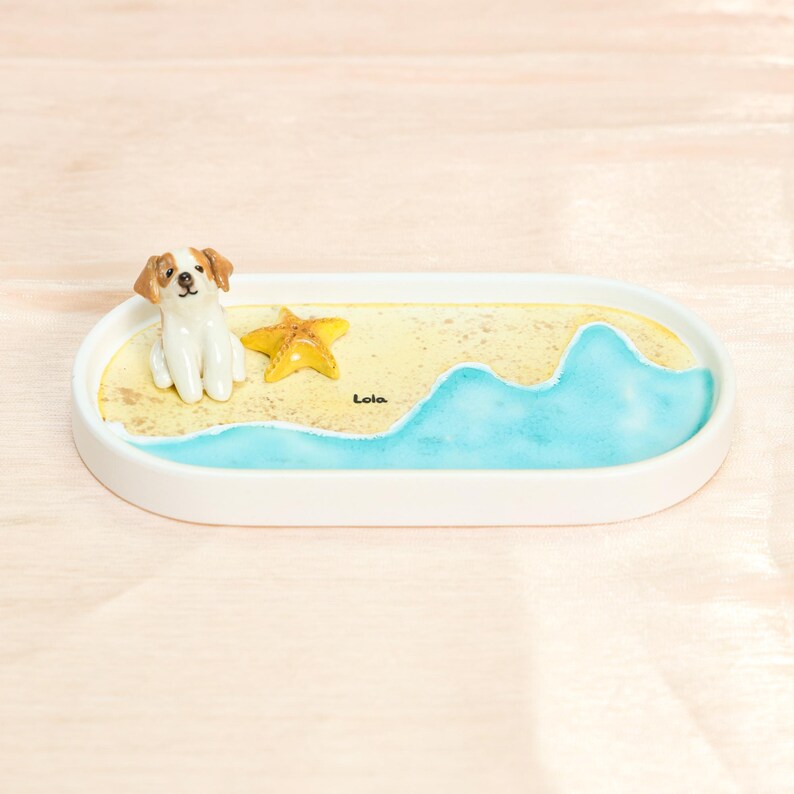 May include: An oval decorative tray featuring a beach scene. A small dog figurine sits on a sandy area beside a yellow starfish. Blue waves and the word "Lola" are painted on the tray.