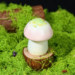 May include: A ceramic mushroom figurine with a pink and green cap and a white stem. The mushroom is set on a small wooden slice, surrounded by green moss. The cap has yellow spots.