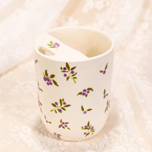 May include: A white ceramic toothbrush holder with a floral design. The holder features a curved top with a slot for a toothbrush and is decorated with purple berry and green leaf motifs.