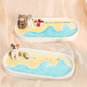 May include: Two ceramic trays with beach scenes. One tray features two cat figurines and a life preserver, the other a dog figurine and a small green object. The trays have a yellow and blue painted design, with the text "Vida y Luna, nuestros dos destinos".