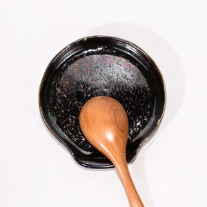 May include: A black ceramic spoon rest with a speckled, iridescent finish. A wooden spoon rests inside the spoon rest. The spoon rest has a curved shape with a raised edge.