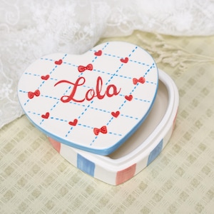 Puede incluir: Caja de baratijas en forma de corazón con tapa. La tapa es blanca con un patrón de cuadrícula, corazones y lazos rojos, y el nombre "Lola" en escritura roja. Los lados de la caja están a rayas azules y rosas.