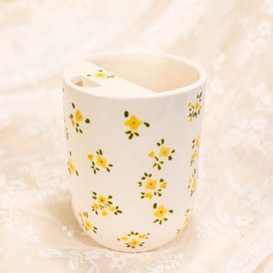 May include: A white ceramic toothbrush holder decorated with a pattern of yellow flowers and green leaves. The holder features a slot for a toothbrush and a rounded form. The floral design is spread across the surface.