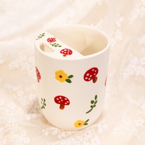 May include: A white ceramic toothbrush holder decorated with red mushroom, yellow flower, and green leaf designs. The holder has a slot for a toothbrush and is a charming addition to any bathroom.