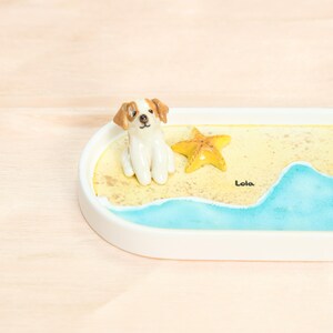 May include: A ceramic tray with a beach scene, featuring a small dog figurine, a yellow starfish, and blue water. The tray has a white border and rounded edges. The name "Lola" is written on the tray. The dog has brown and white markings.