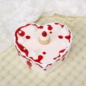 May include: A heart-shaped ceramic trinket box with a pink surface, decorated with red drizzles and white accents. It features a small, decorative cake-like element on top. The box is on a patterned surface.