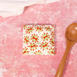 Puede incluir: Un plato cuadrado de cerámica con bordes festoneados que presenta un patrón repetitivo de pequeños lazos rojos rodeados de diseños de rayos de sol en verde claro, acompañado de una cuchara de madera lisa con cuenco redondo y mango largo.