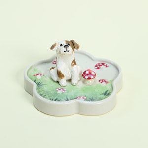 May include: A ceramic figurine of a dog with brown and white markings sits on a cloud-shaped base. The base features a painted green lawn with red and white mushroom accents. The figurine is a decorative item.